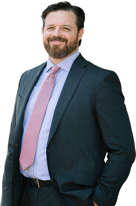 A man with dark hair and a beard, wearing a dark striped suit, light blue shirt, and pink tie, stands smiling with one hand in his pocket. A man with dark hair and a beard, wearing a dark striped suit, light blue shirt, and pink tie, stands smiling with one hand in his pocket.