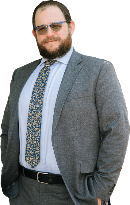 A man with dark hair and a beard, wearing a dark striped suit, light blue shirt, and pink tie, stands smiling with one hand in his pocket. A man with dark hair and a beard, wearing a dark striped suit, light blue shirt, and pink tie, stands smiling with one hand in his pocket.