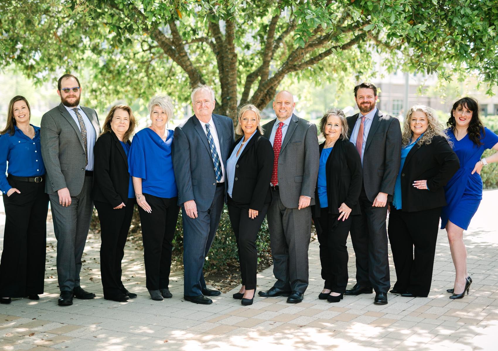 A group of eleven professionally dressed adults stands outdoors in front of a leafy tree, smiling at the camera. Most are wearing business attire in shades of blue, black, and gray. A group of eleven professionally dressed adults stands outdoors in front of a leafy tree, smiling at the camera. Most are wearing business attire in shades of blue, black, and gray.