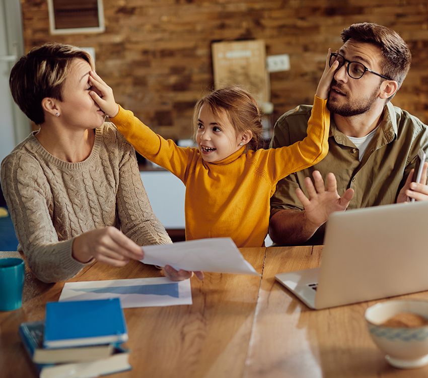 A smiling child in a yellow top playfully covers the eyes of both adults sitting beside her at a table with papers, books, and a laptop. The adults react with amusement in a cozy home setting.
