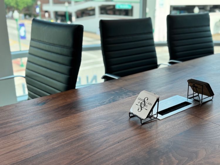 A close-up of a wooden conference table with black leather chairs, business cards on display stands, and large windows overlooking a street outside. A close-up of a wooden conference table with black leather chairs, business cards on display stands, and large windows overlooking a street outside.
