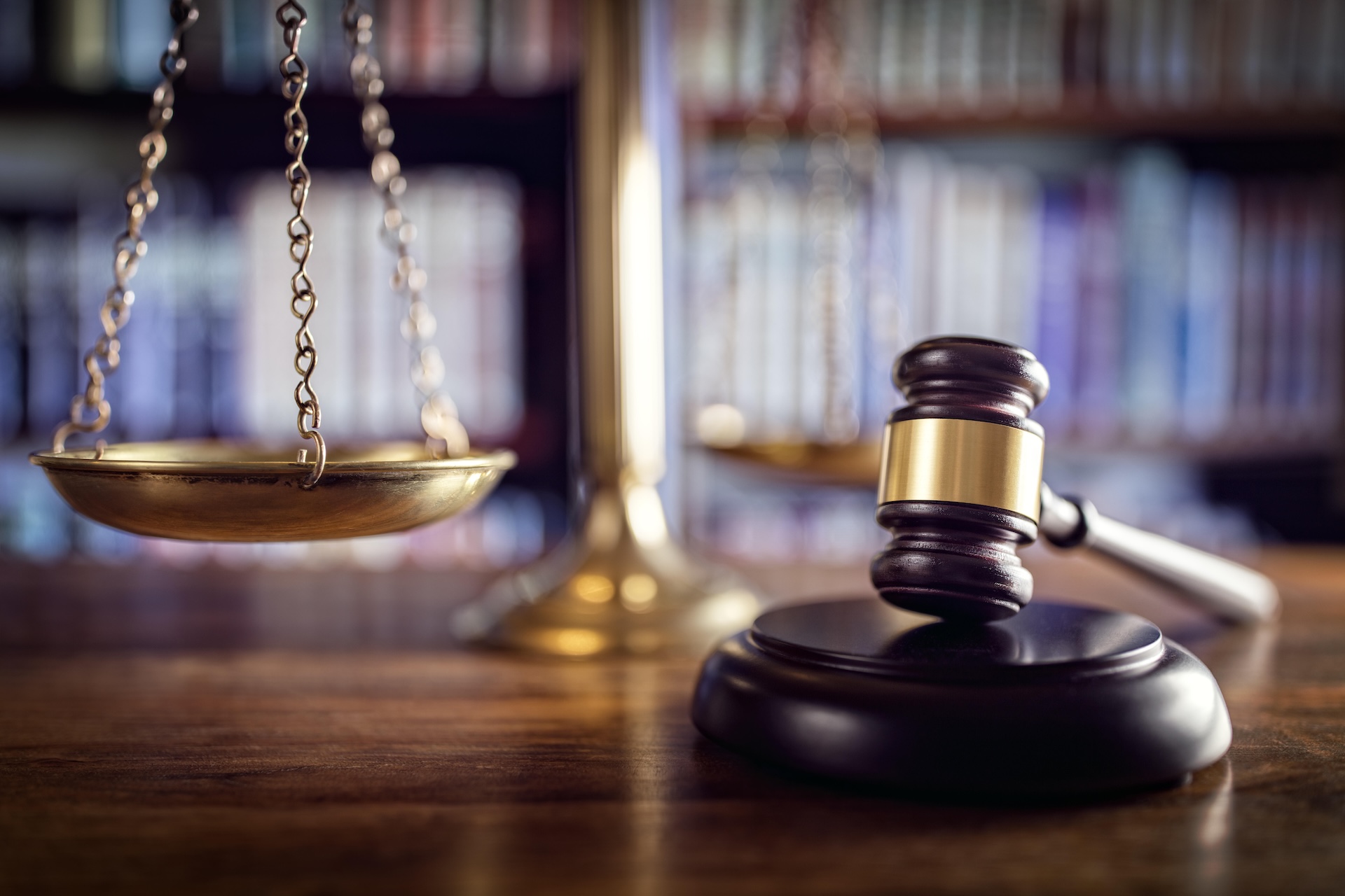 A close-up of a judge’s gavel resting on a sound block next to brass balance scales, with blurred bookshelves in the background, symbolizing law and justice.