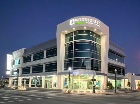 Modern three-story Woodforest National Bank building at dusk, with large glass windows and illuminated signage, located at a street corner with visible streetlights and traffic signals. Modern three-story Woodforest National Bank building at dusk, with large glass windows and illuminated signage, located at a street corner with visible streetlights and traffic signals.
