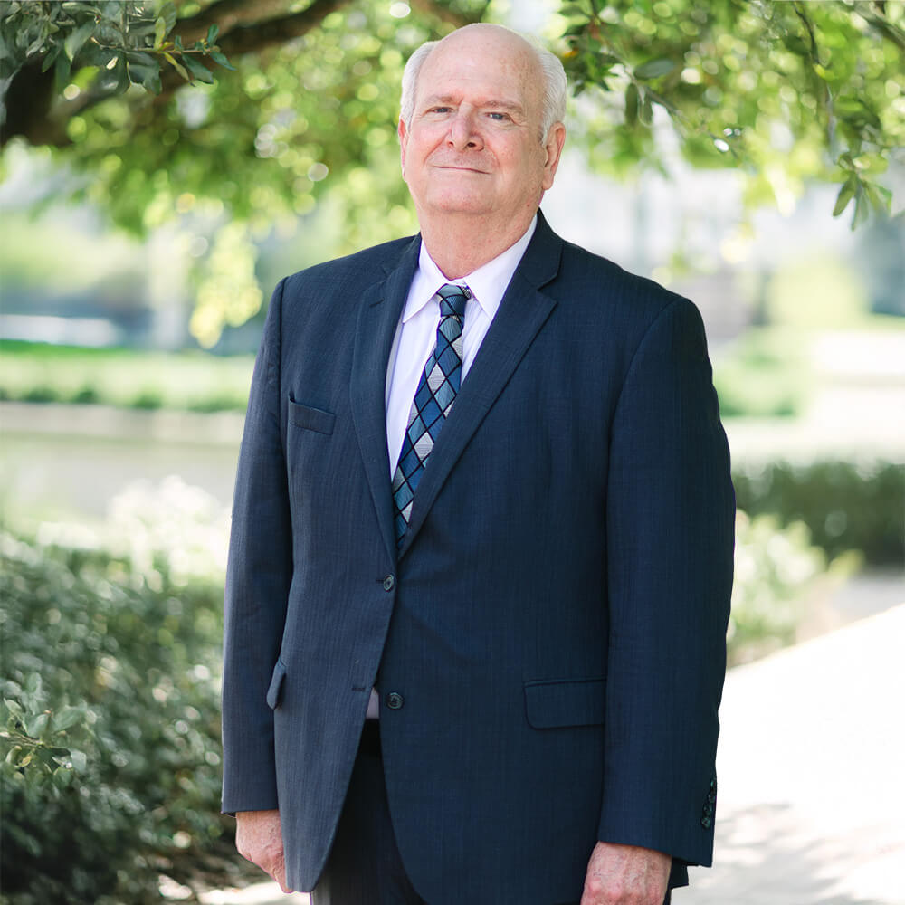 An older man with short white hair wearing a dark suit, light shirt, and patterned tie stands outside in a sunlit garden, with trees and greenery in the background.