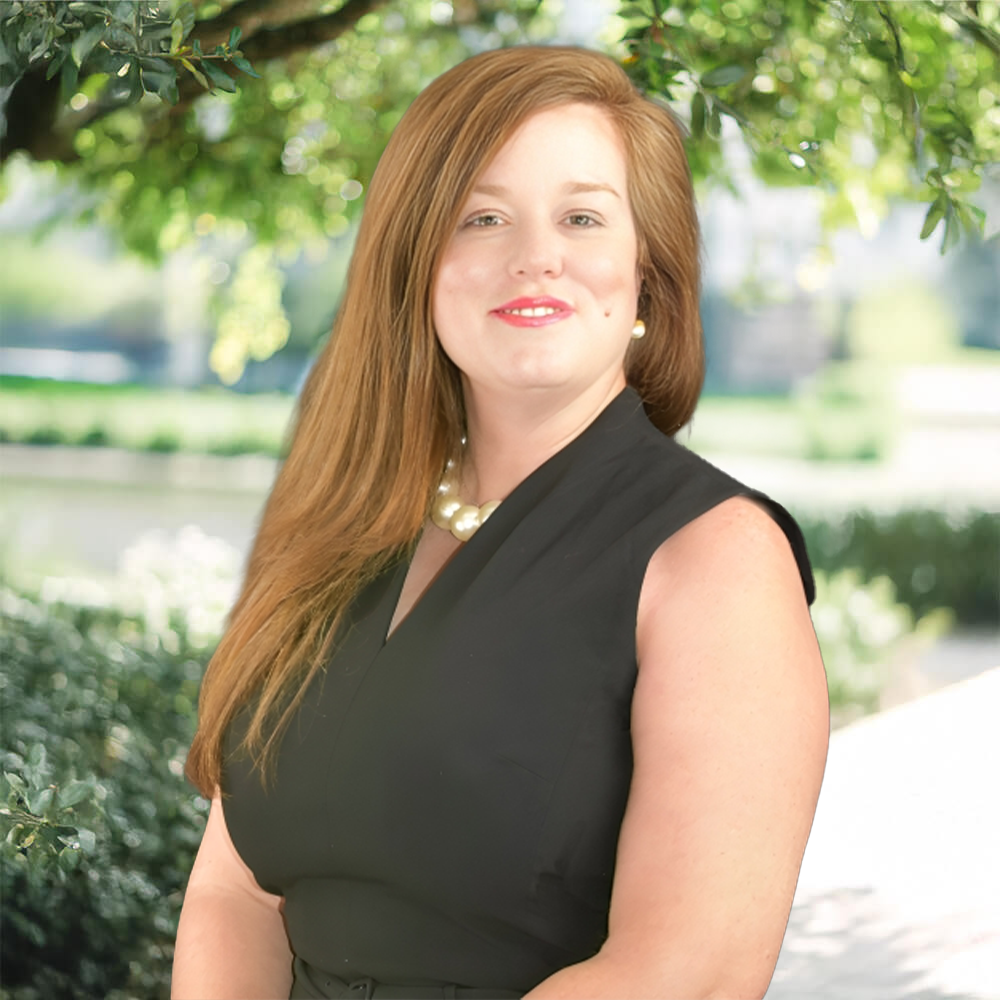 A woman with long reddish-brown hair, wearing a sleeveless black dress and a pearl necklace, is smiling outdoors with green trees and bright sunlight in the background.