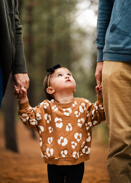 A young child wearing a brown patterned sweatshirt and a black bow looks up while holding hands with two adults, standing between them outdoors in a wooded area.
