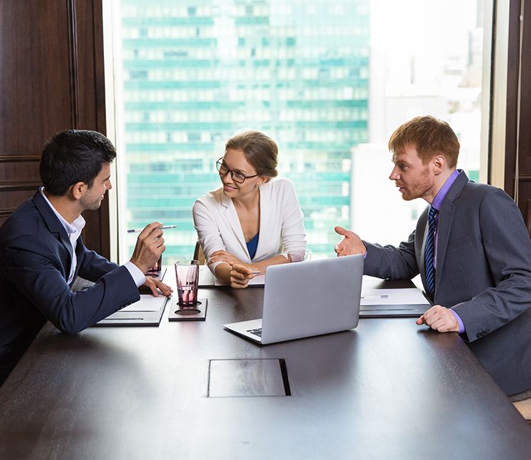 Three business professionals sit around a table in an office, engaged in a discussion. Two men and one woman are talking, with a laptop and documents in front of them. A large window shows a cityscape in the background. Three business professionals sit around a table in an office, engaged in a discussion. Two men and one woman are talking, with a laptop and documents in front of them. A large window shows a cityscape in the background.