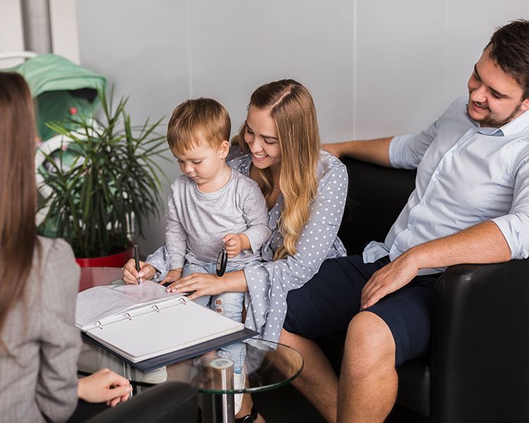 A smiling family sits together in an office. A woman helps a young child hold a pen over paperwork on a glass table, while a man sits beside them and another person faces them across the table.
