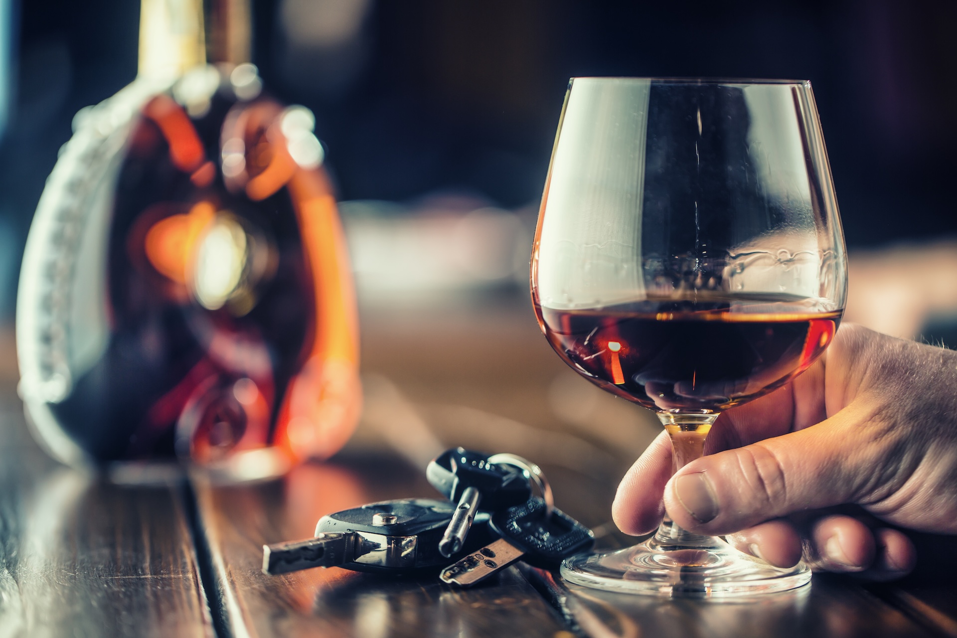 A hand holding a glass of amber alcohol sits next to car keys on a wooden table, with a blurred liquor bottle in the background, suggesting the risks of drinking and driving. A hand holding a glass of amber alcohol sits next to car keys on a wooden table, with a blurred liquor bottle in the background, suggesting the risks of drinking and driving.