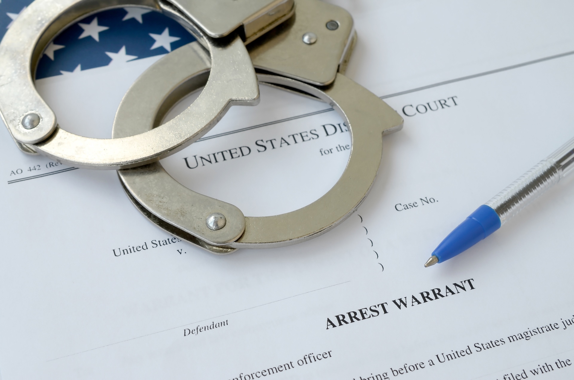 A pair of handcuffs and a blue pen rest on an official document labeled "Arrest Warrant" from a United States District Court, with part of an American flag visible in the background.