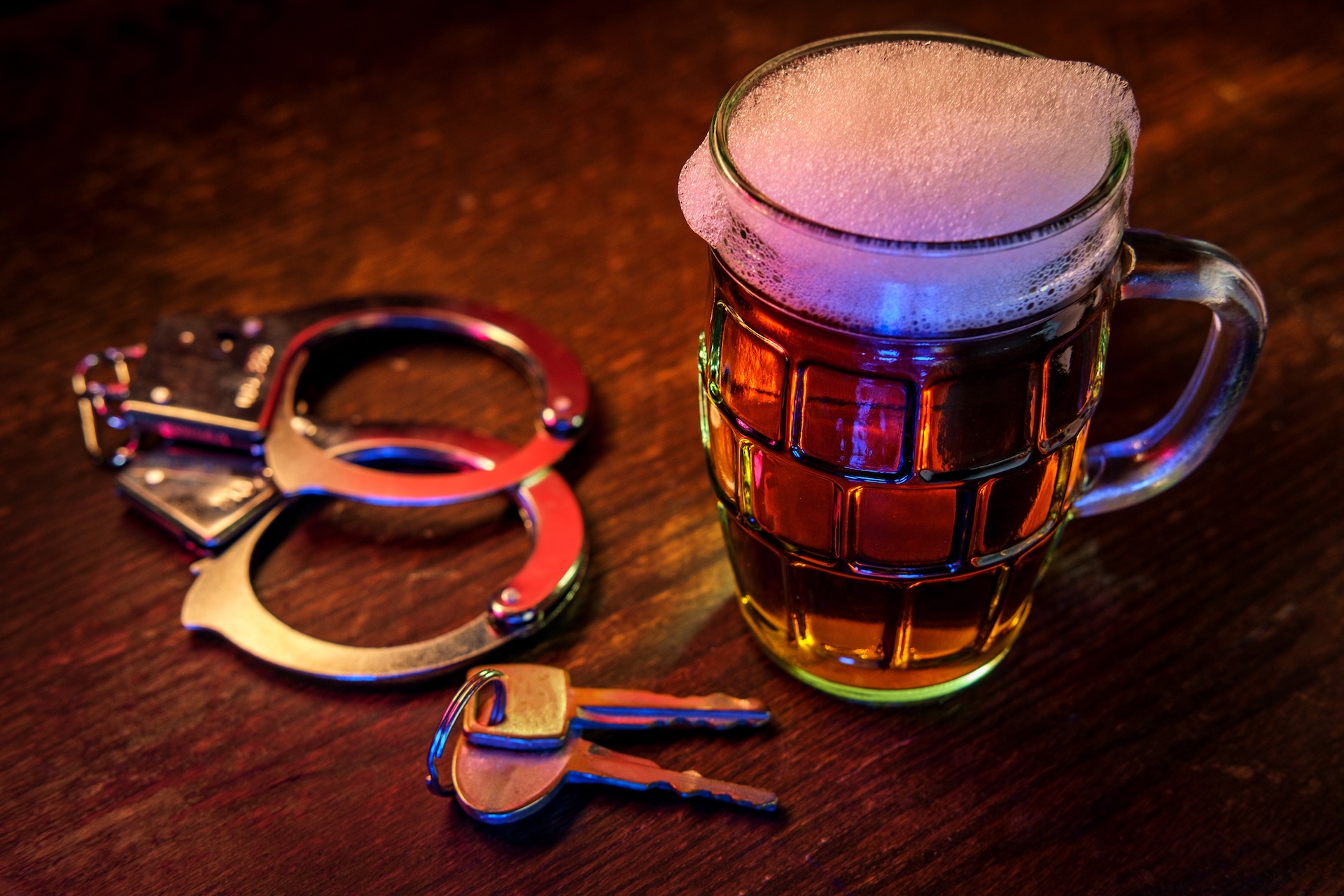 A frothy beer mug sits on a wooden surface beside a set of handcuffs and car keys, illuminated by colorful light, symbolizing the dangers of drinking and driving.