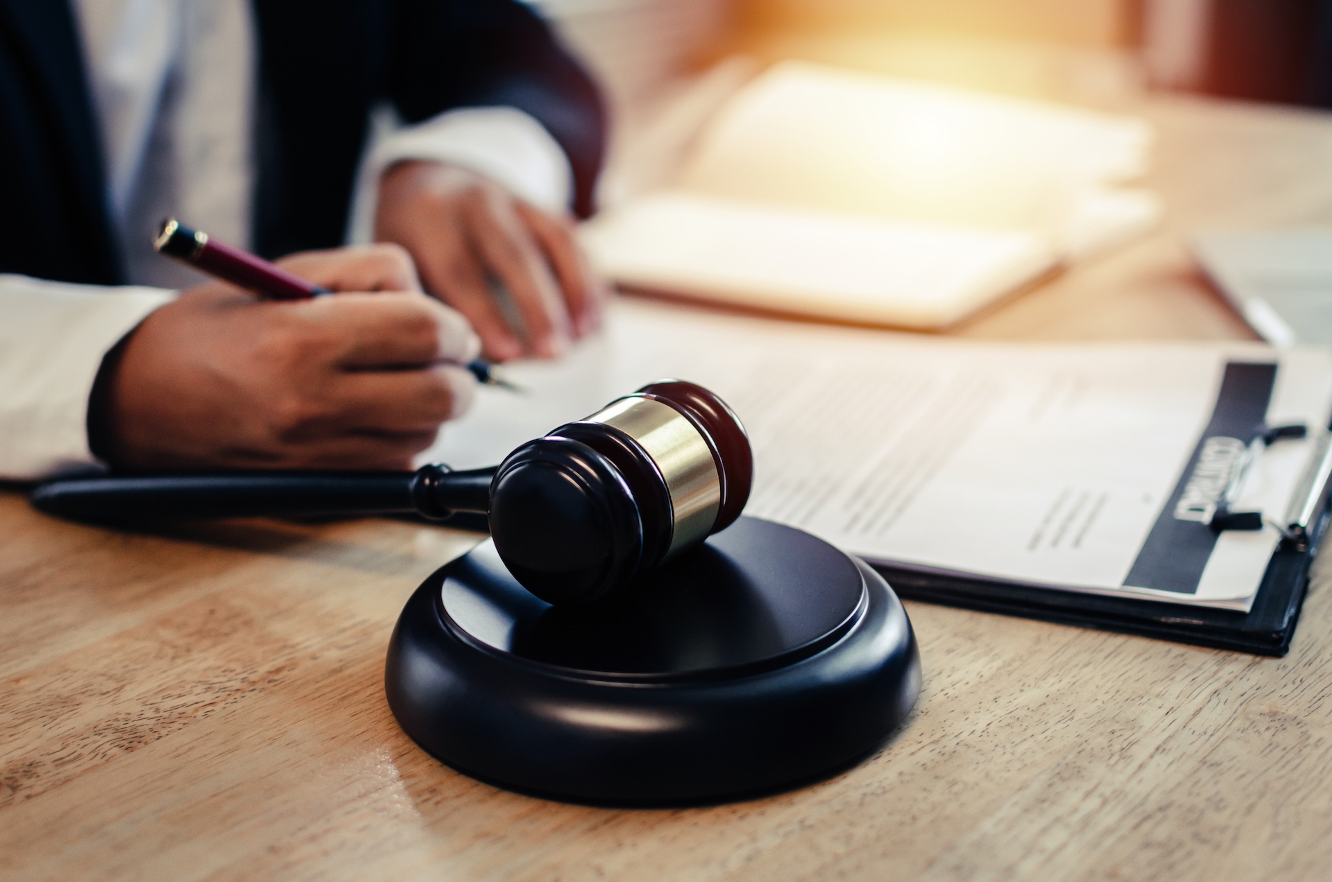 A person in a suit writes on a document at a desk with a gavel and legal paperwork in the foreground, suggesting a legal or judicial setting.