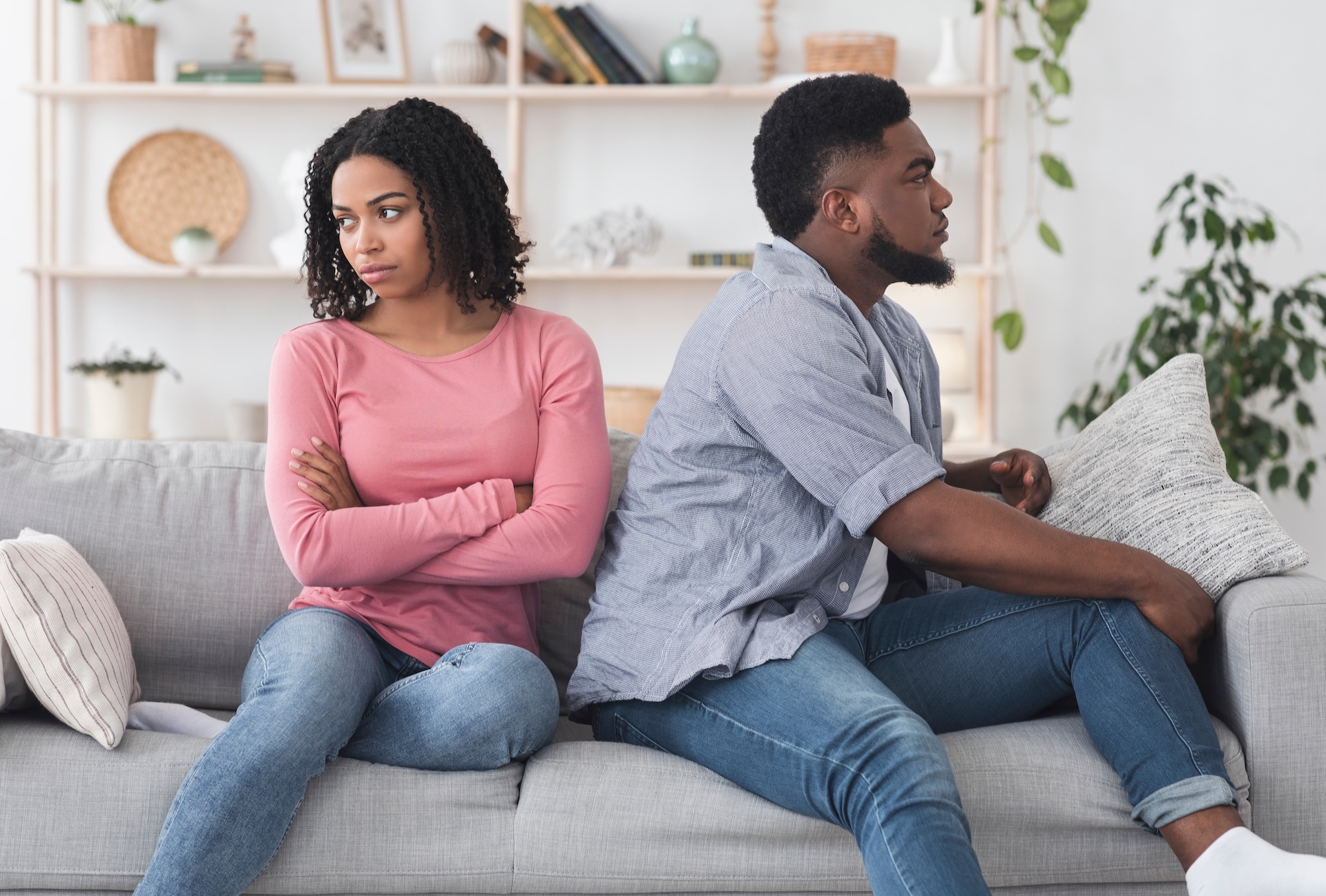 A woman and a man sit back-to-back on a couch, both looking away from each other with serious expressions. The woman has her arms crossed, and the atmosphere appears tense or uncomfortable.