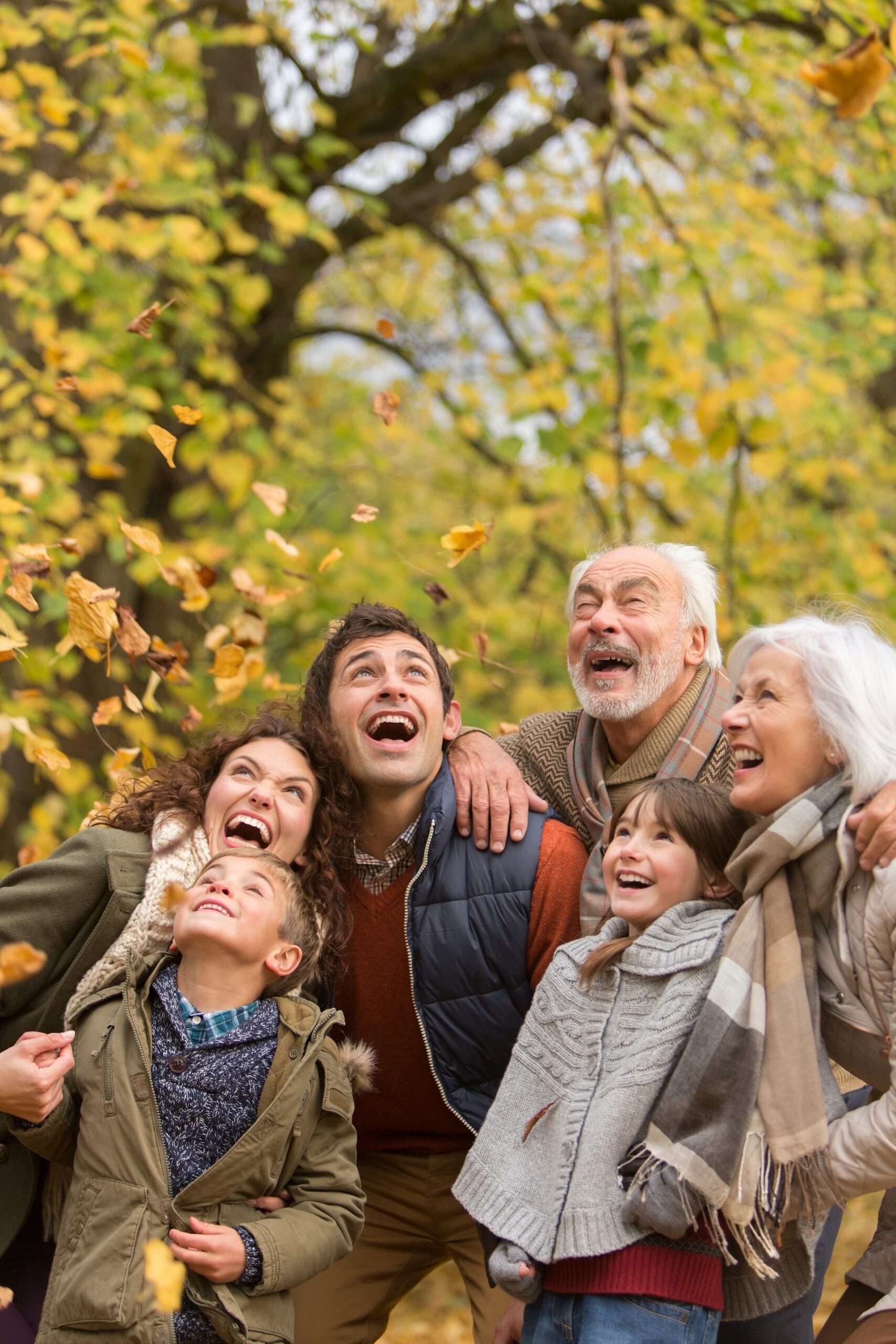 A smiling family sits together in an office. A woman helps a young child hold a pen over paperwork on a glass table, while a man sits beside them and another person faces them across the table.