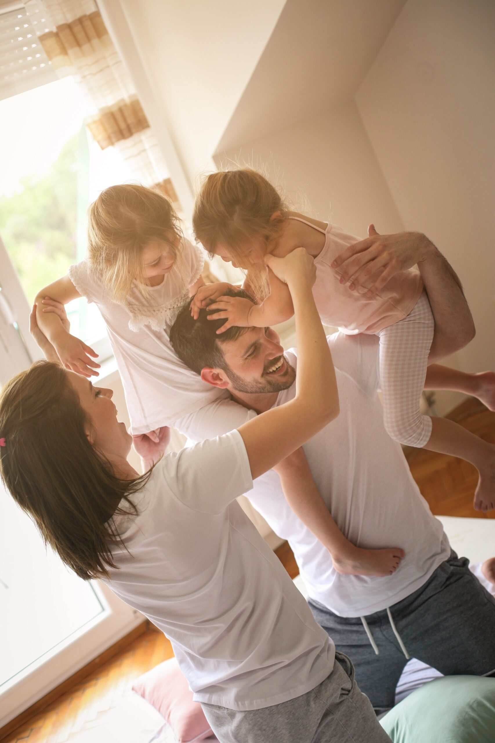 Cheerful family playing together on bed. Parents spending free time with their daughters. Daughters sitting on fathers shoulders. A smiling child in a yellow top playfully covers the eyes of both adults sitting beside her at a table with papers, books, and a laptop. The adults react with amusement in a cozy home setting.