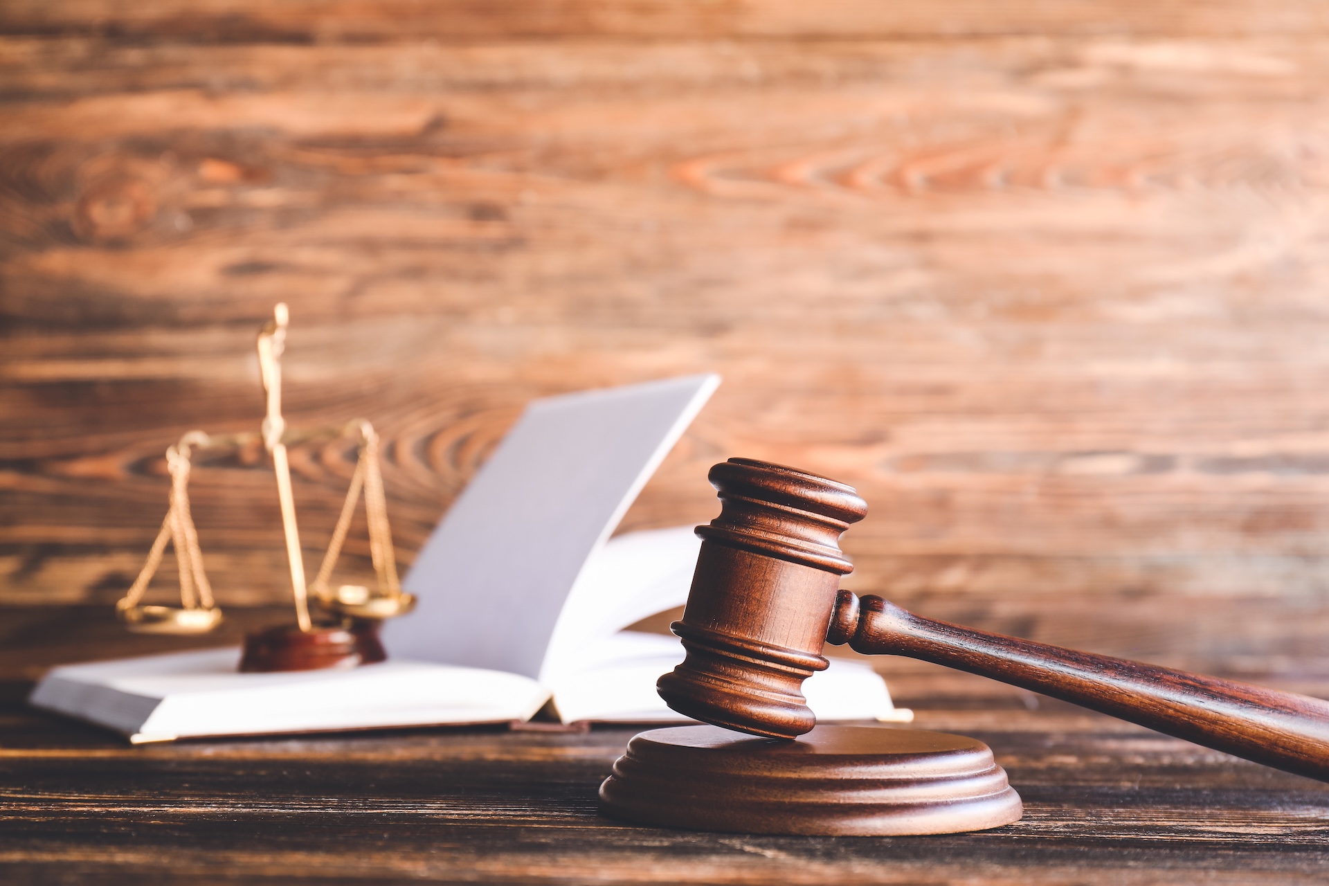 A wooden judge's gavel rests on a sound block in the foreground, with a balance scale and an open book in the blurred background, all set on a wooden surface.