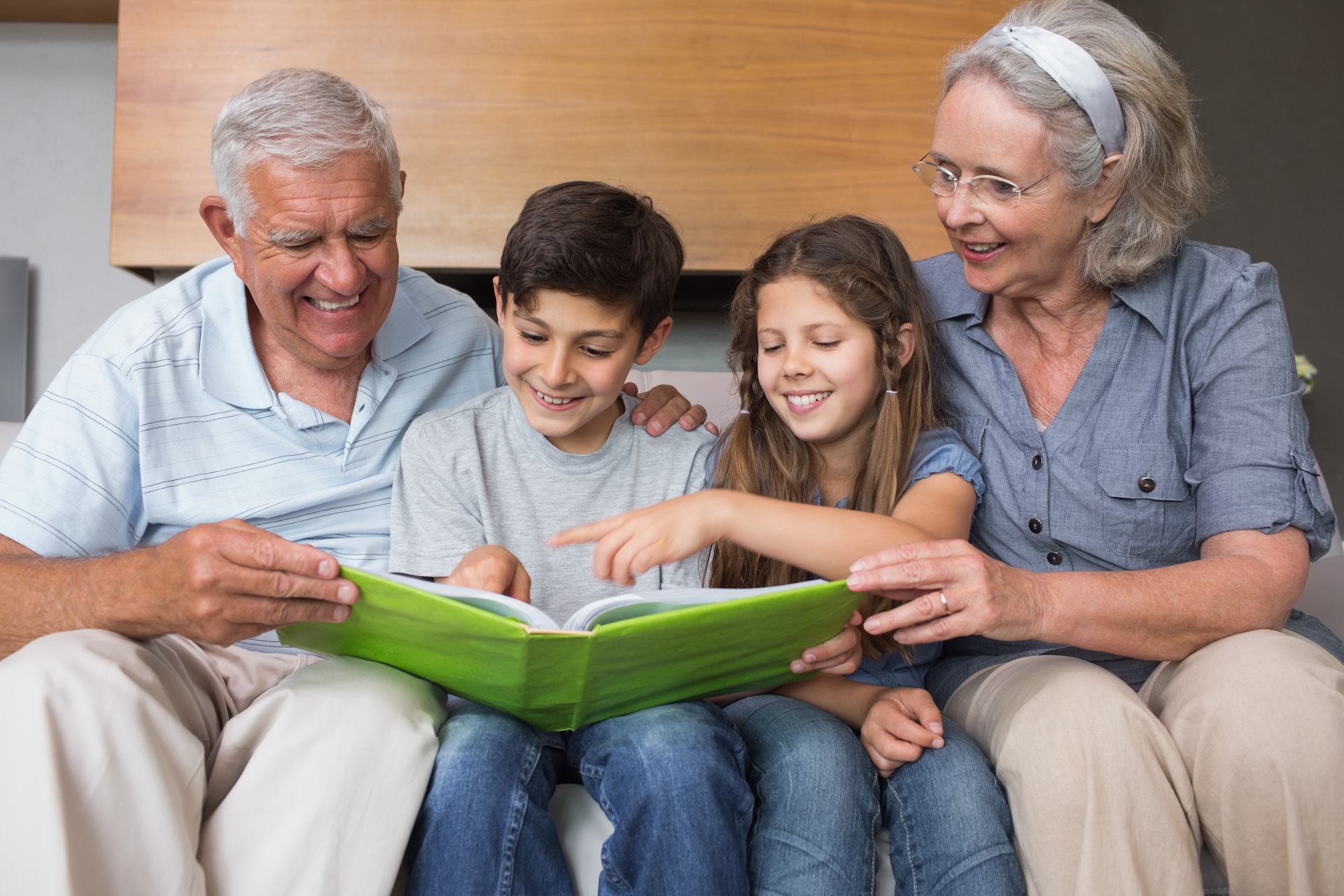 An elderly couple sits on a couch with a young boy and girl, all smiling as they look at and point to a large green book together.