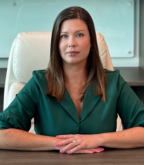 A woman with straight brown hair, wearing a green blouse, sits at a desk with her hands folded. She has a neutral expression and is seated in a white office chair with a light background behind her.
