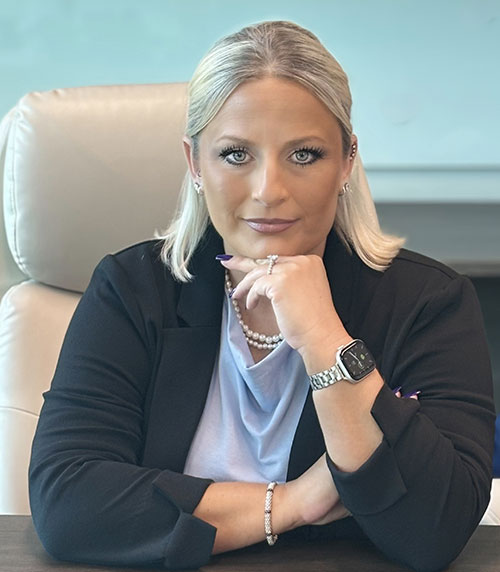 A woman with blonde hair, wearing a black blazer, light top, and pearl jewelry, sits at a desk resting her chin on her hand. She looks directly at the camera with a confident expression.