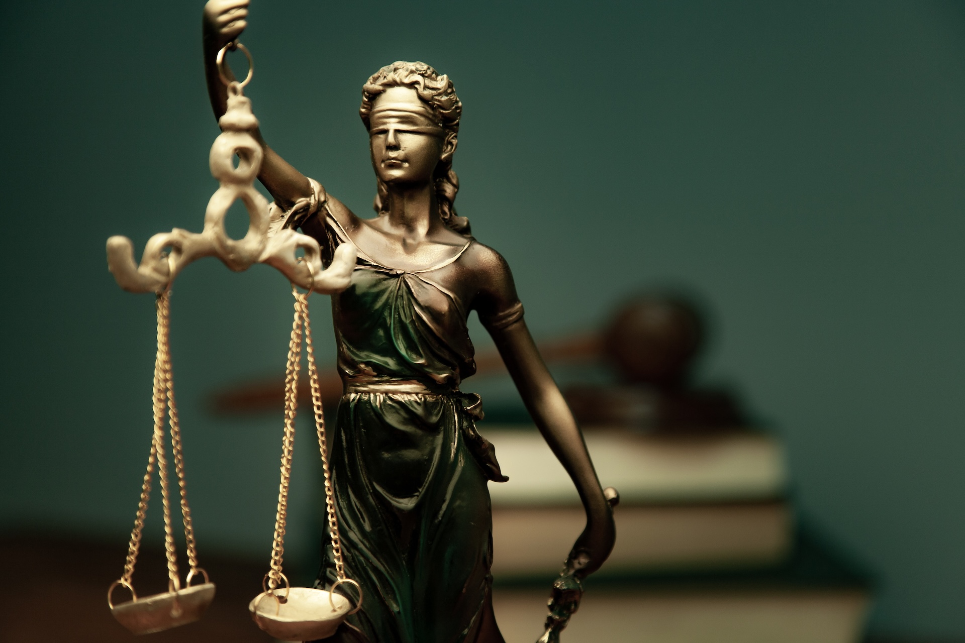 A close-up of a bronze statue of Lady Justice holding scales, wearing a blindfold. In the blurred background, a judge’s gavel rests on stacked books, symbolizing law and justice. A close-up of a bronze statue of Lady Justice holding scales, wearing a blindfold. In the blurred background, a judge’s gavel rests on stacked books, symbolizing law and justice.