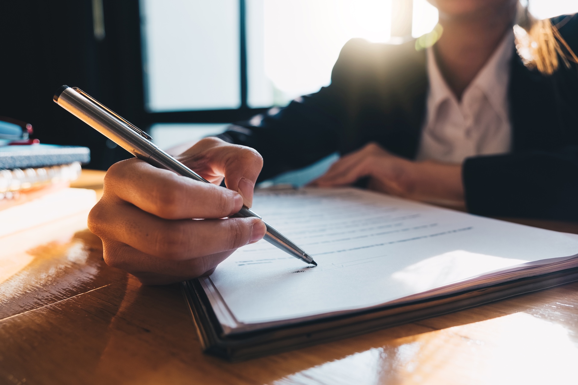 A person in business attire sits at a desk, holding a pen and signing a document, with sunlight streaming through a window in the background. A person in business attire sits at a desk, holding a pen and signing a document, with sunlight streaming through a window in the background.