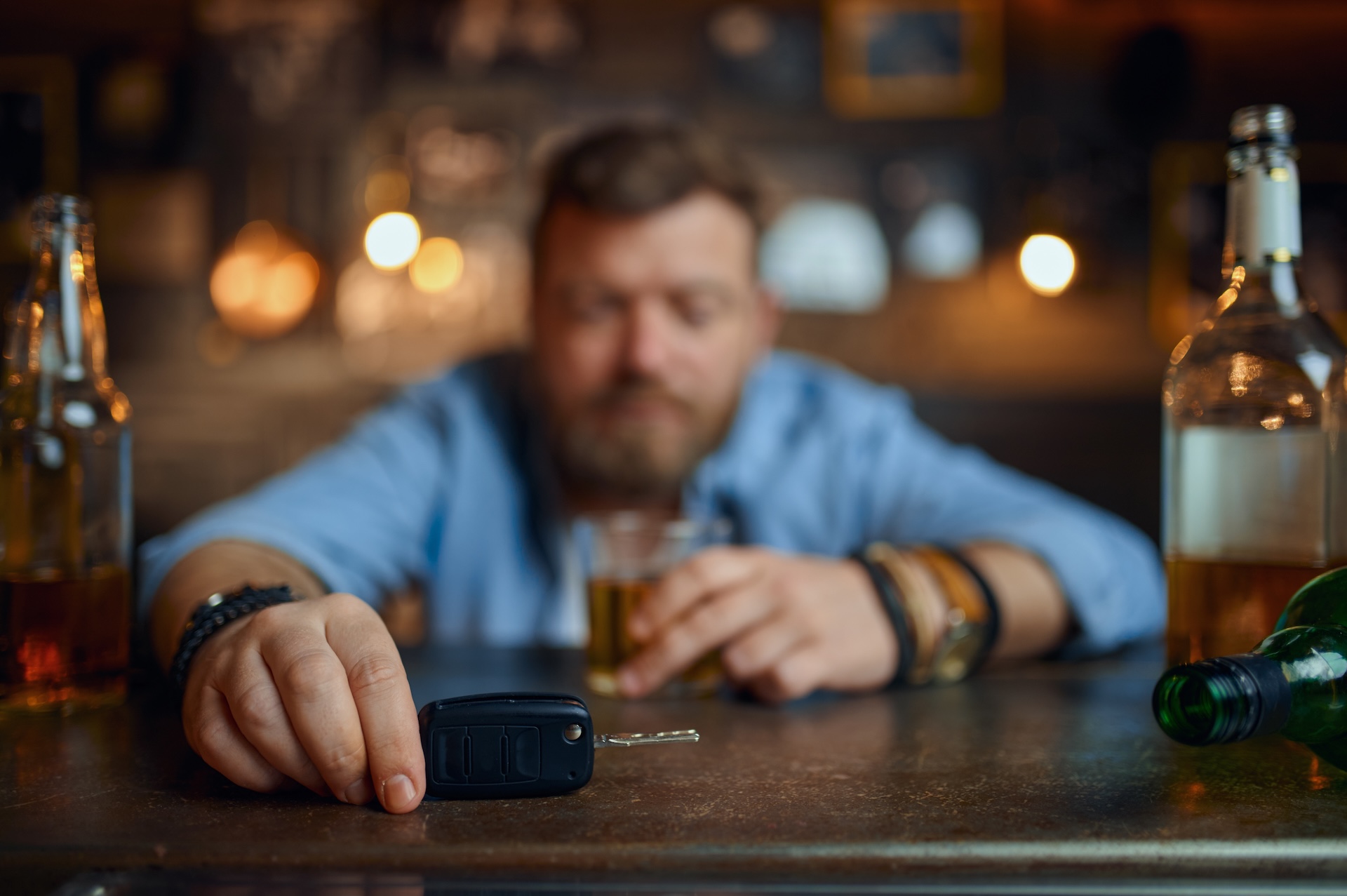 A man with a beard sits at a bar holding a glass of alcohol, with several bottles around him. His other hand rests on a set of car keys on the counter, suggesting the dangers of drinking and driving. A man with a beard sits at a bar holding a glass of alcohol, with several bottles around him. His other hand rests on a set of car keys on the counter, suggesting the dangers of drinking and driving.