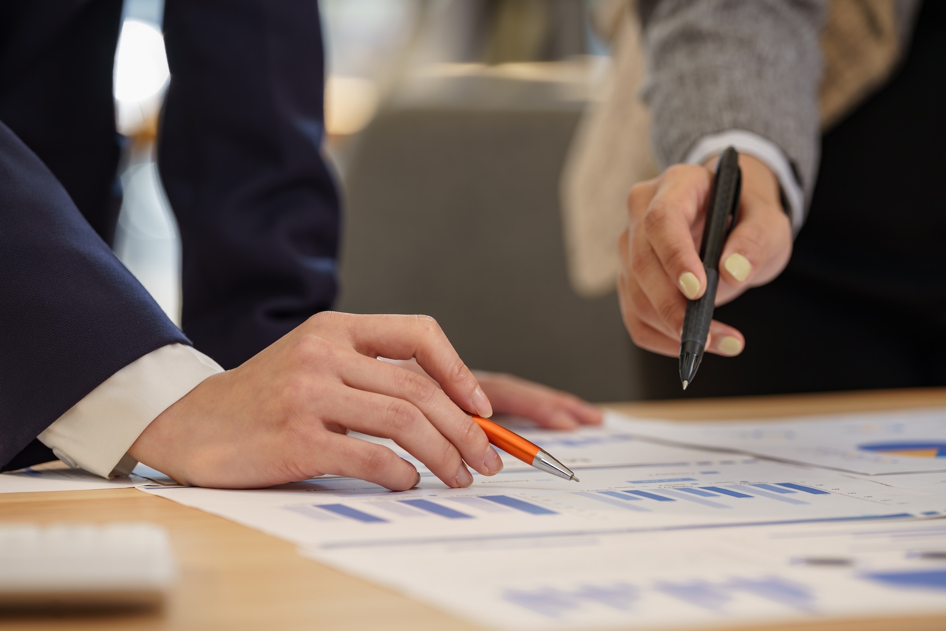 Two people reviewing printed charts and graphs on a table, one holding an orange pen and the other pointing with a black pen, suggesting discussion or analysis of financial or business data. Two people reviewing printed charts and graphs on a table, one holding an orange pen and the other pointing with a black pen, suggesting discussion or analysis of financial or business data.