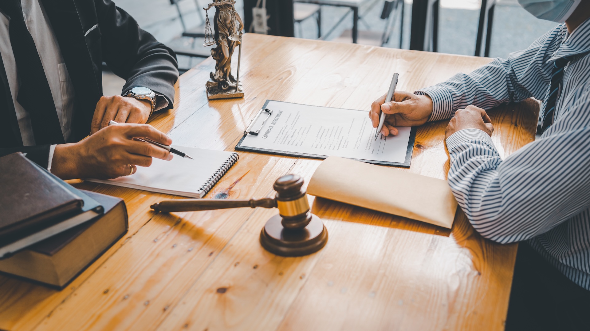 Two people sit at a wooden table with legal documents, a gavel, law books, and a small Lady Justice statue, suggesting a legal consultation or meeting. One person points while the other writes on a clipboard. Two people sit at a wooden table with legal documents, a gavel, law books, and a small Lady Justice statue, suggesting a legal consultation or meeting. One person points while the other writes on a clipboard.